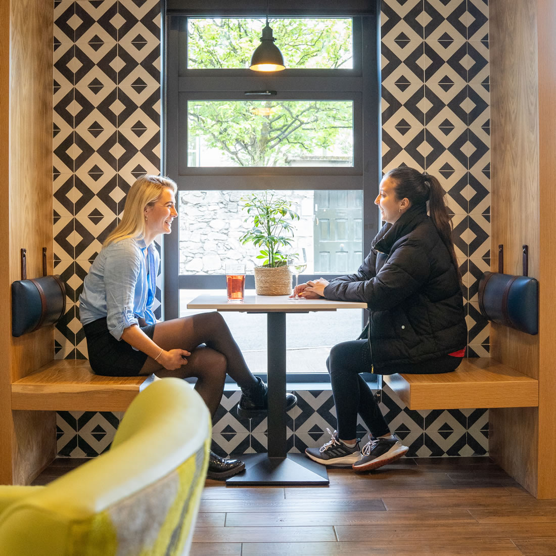 Two friends enjoying drinks and conversation in a stylish window booth at Snoozles Hostel common area